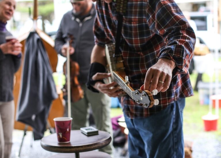 A person in a plaid shirt tunes a banjo while others stand nearby, one holding a violin. Musicians at the Market gather outdoors as a red paper cup sits on a round table in the foreground.