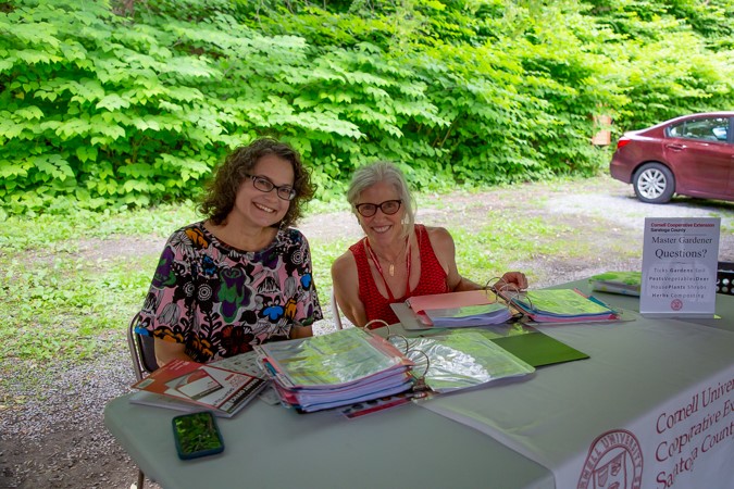 Two smiling women from a local non-profit sit at an outdoor table covered with binders, papers, and a phone. A sign offers “Master Gardener Questions?” Leafy green bushes and a red car are visible in the background.