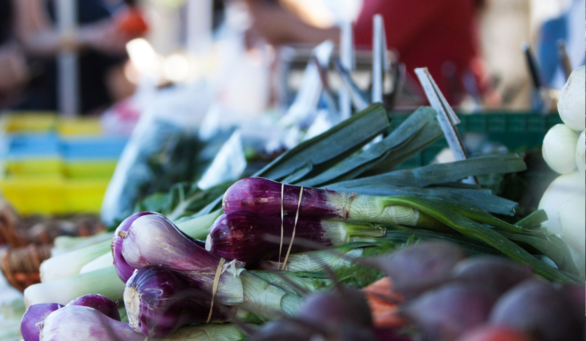 Bundles of fresh green onions and purple vegetables are displayed on a market table at Clifton Park Market, with blurred shoppers and other produce in the background.