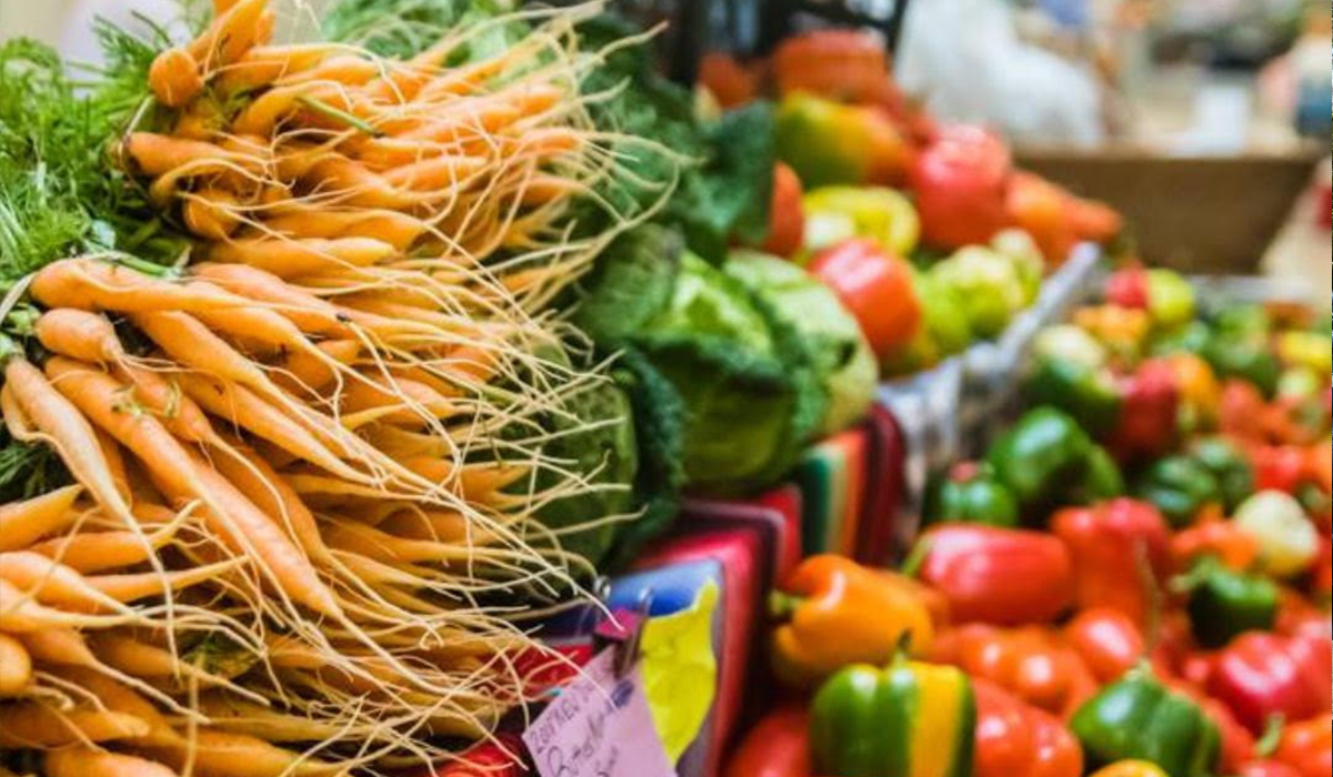 A colorful assortment of fresh vegetables at a winter market, including bunches of carrots with long roots, green cabbage, and various bell peppers in yellow, red, and green hues.