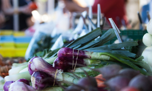 Clifton-Park-Market Bundles of fresh green onions and purple vegetables are displayed on a market table at Clifton Park Market, with blurred shoppers and other produce in the background.