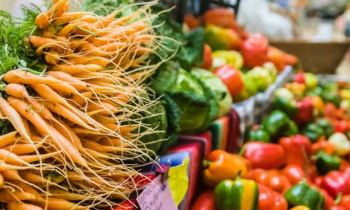 inside-market-1 A colorful assortment of fresh vegetables at a winter market, including bunches of carrots with long roots, green cabbage, and various bell peppers in yellow, red, and green hues.
