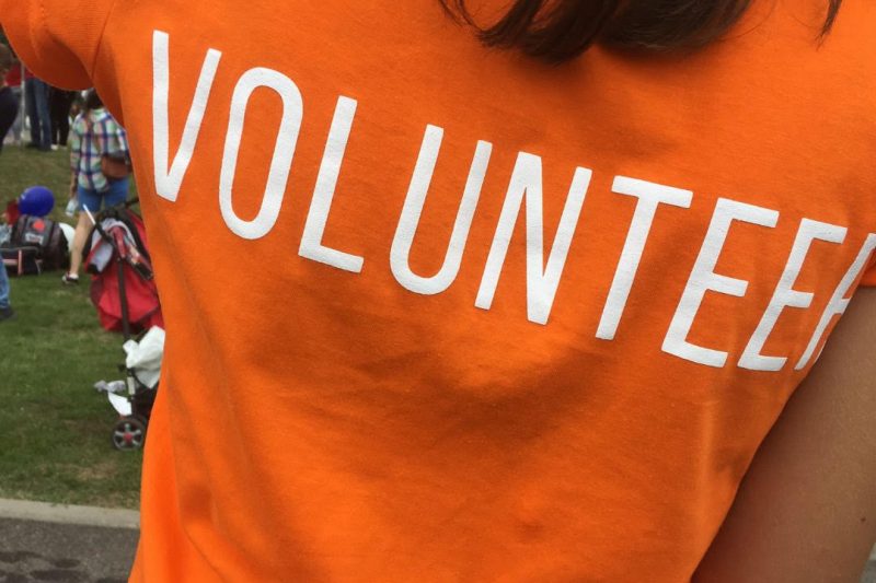 volunteer A person wearing an orange shirt with the word VOLUNTEER printed in large white letters on the back stands outdoors, inspiring others to get involved.